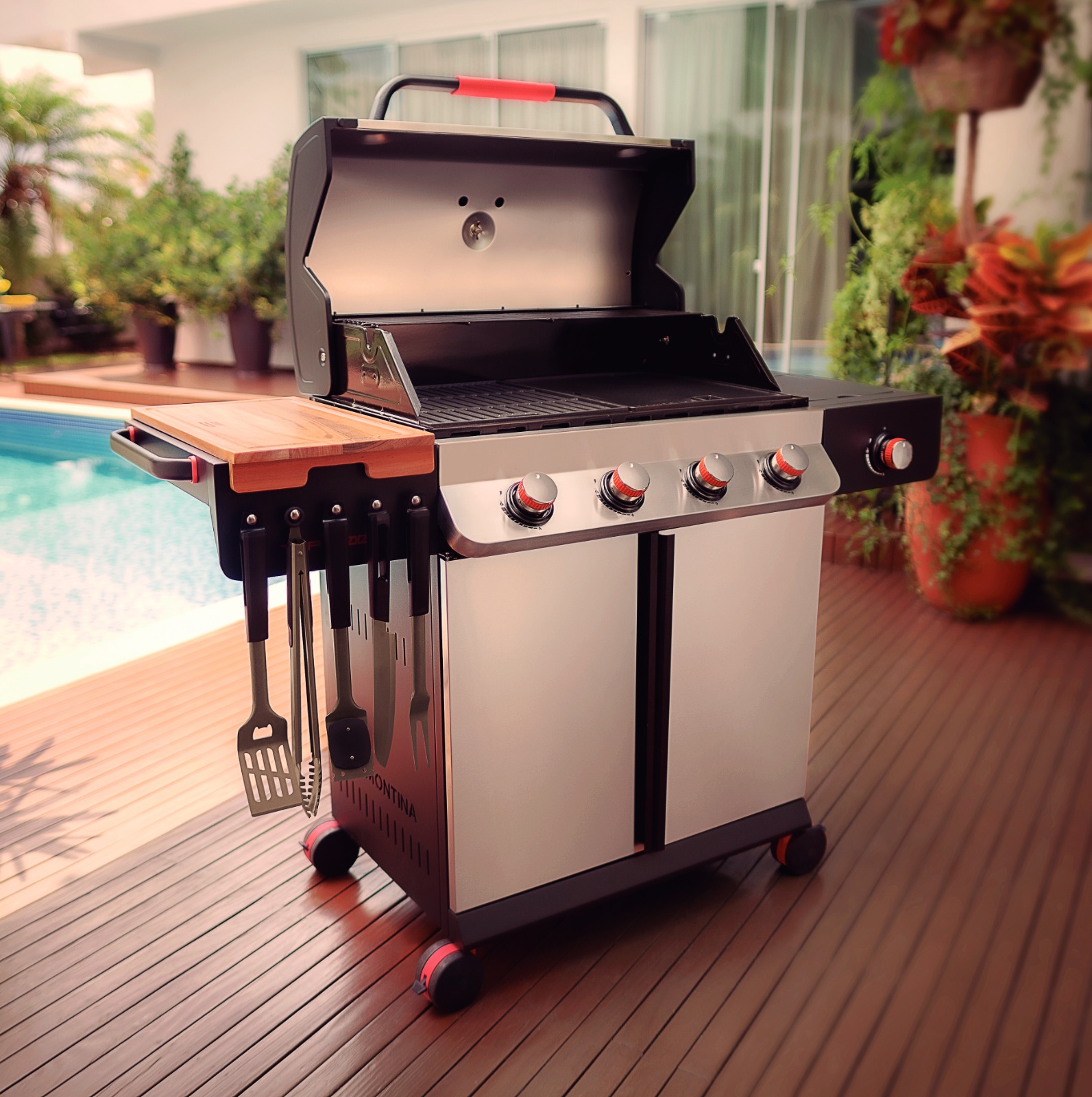 Churrasqueira a gás Tramontina TGP 6000 em área externa, com utensílios pendurados e tampo de madeira ao lado de piscina.